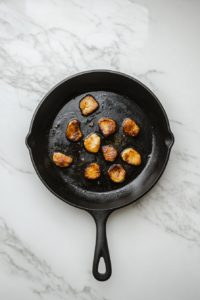 This image shows golden brown chicken pieces cooking in melted butter inside a black cast-iron skillet on a white marble countertop.