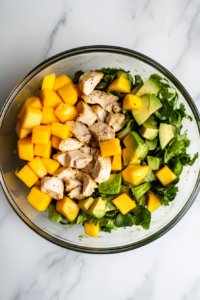 This image shows a clear glass bowl sitting on a white marble countertop filled with tender cooked chicken chunks, golden mango pieces, and ripe avocado cubes, freshly tossed together.