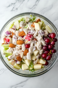 This image shows all the chicken salad ingredients fully coated with the creamy dressing in a large clear glass bowl on a white marble countertop.