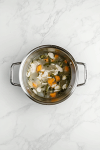 This image shows chicken stock poured over lightly browned vegetables in a stainless steel pot placed on a white marble cooktop.