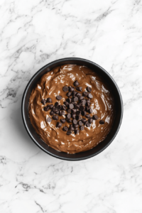 This image shows the baked chocolate chocolate chip cake cooling inside a black Bundt pan on a wire rack over a white marble countertop.