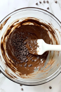This image shows semisweet chocolate chips stirred into smooth chocolate cake batter inside a clear glass bowl over a white marble countertop.