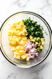 This image shows creamy avocado chunks, finely chopped onions, mayonnaise, pickles, and mustard being added to the mashed egg mixture.