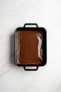 This image shows a black baking dish filled with evenly spread cinnamon roll cake batter on a clean white marble surface.