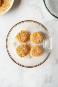 This image shows chicken patties being coated with breadcrumbs in a shallow dish, on a clean white marble surface.