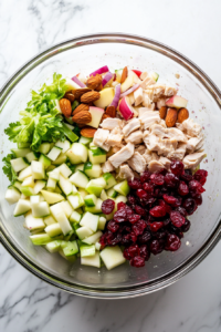 This image shows cooked chicken, chopped celery, diced red onion, halved red grapes, chopped apple, dried cranberries, and slivered almonds tossed in a large clear glass mixing bowl on a white marble countertop.