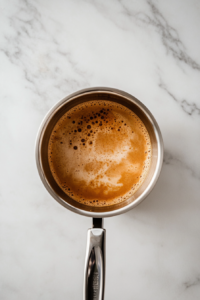 This image shows caramel bubbling in a stainless steel saucepan with a thermometer inserted, cooking over a clean white marble cooktop.