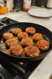 This image shows chicken patties cooking in olive oil in a black skillet, browning on one side, over a white marble surface.