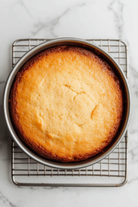 This image shows the freshly baked yogurt cake still in the round pan, resting on a wire rack set on a white marble cooktop, cooling for ten minutes before removal.