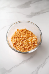 This image shows crushed crackers and melted butter mixed in a small clear glass bowl on a white marble countertop.