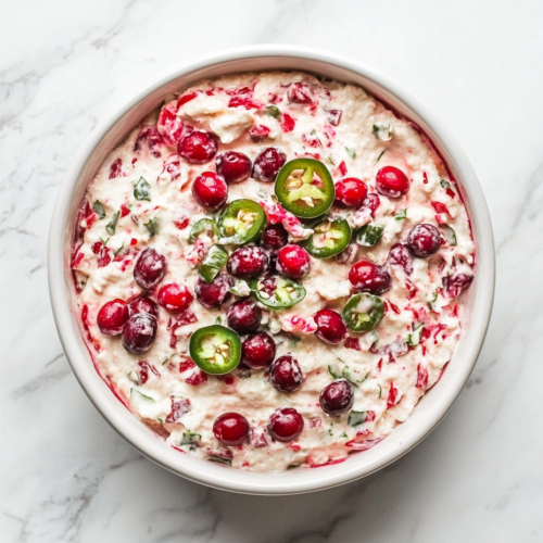 This image shows a creamy cranberry jalapeño dip served in a white round bowl, with a vibrant pink hue and topped with finely chopped cranberries and jalapeños for a festive and zesty look.