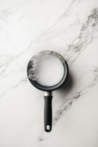 This image shows heavy cream heating until steaming in a small black nonstick saucepan on a white marble countertop.