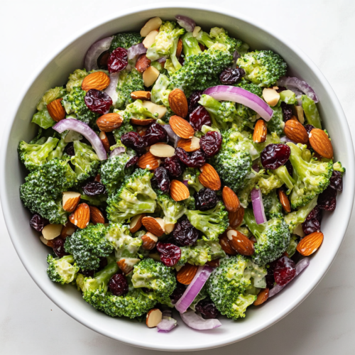 This image shows a vibrant broccoli salad served in a round white bowl, infused with fresh broccoli florets, sliced onions, creamy dressing, sweet raisins, and crunchy almonds.