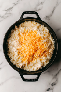 This image shows a creamy mixture of rice, chicken, cream of chicken soup, milk, sour cream, seasoning, and shredded cheddar cheese in a black skillet on a white marble countertop.