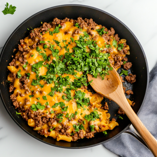 This image shows a bowl of creamy cheeseburger pasta topped with melted cheddar cheese and a sprinkle of fresh parsley, served in a white ceramic bowl on a clean white marble countertop.