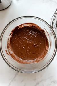 This image shows a smooth, creamy chocolate cake batter inside a clear glass mixing bowl placed on a white marble countertop.