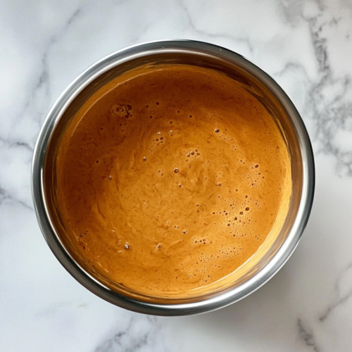 This image shows a top-down view of creamy, melted cheese sauce in a stainless steel saucepan resting on a clean white marble cooktop with no other elements in the background.