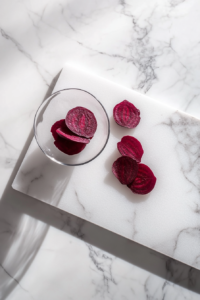 This image shows hands peeling and slicing deep red beetroot on a wooden cutting board, with beet juice staining the board slightly and slices fanned out neatly beside a peele