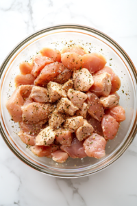 This image shows raw diced chicken seasoned with Italian seasoning, salt, and pepper in a large clear glass bowl on a white marble countertop.