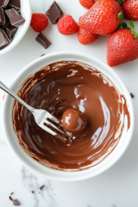 This image shows a chilled Butterfinger ball being dipped into melted chocolate using a silver fork. Some balls are already coated and placed on the parchment-lined baking sheet, which rests neatly on the white marble cooktop, capturing every stage of the chocolate coating process.