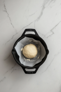 Top-down view of unbaked dough placed on parchment paper inside a black cast iron hotpot on a clean white marble countertop.
