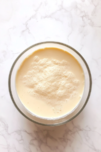 This image shows flour, sugar, baking powder, eggs, vanilla, and milk gathered in a clear glass bowl on a white marble countertop.