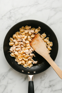 This image shows dry Banza pasta shells being stirred into a bubbling skillet of seasoned chicken broth and ground chicken on a white marble cooktop.