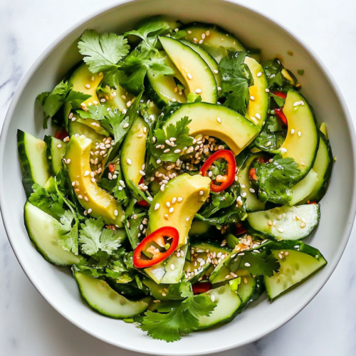 This image shows a beautifully plated Asian Avocado Salad in a round bowl, featuring vibrant avocado slices, cucumber, and fresh herbs topped with white sesame seeds.