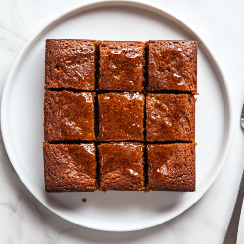 This image shows a traditional Parkin cake, dark brown in color with a glossy glazed surface, cut into squares and neatly arranged on a round white plate.
