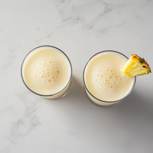 Two light yellow pina coladas are served in cocktail glasses, each garnished with a fresh pineapple slice, giving a tropical and refreshing vibe.