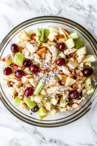 This image shows the finished chicken salad in a clear glass bowl on a white marble countertop, with visible flecks of salt and pepper freshly sprinkled on top
