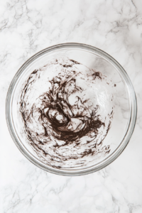 This image shows chocolate batter being gently folded into whipped egg whites in a glass bowl over a white marble countertop.