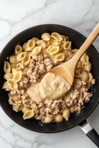This image shows shredded cheddar cheese being folded into the cooked cheeseburger pasta and sauce mixture in a black skillet on the white marble cooktop.