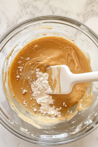 This image shows the dry ingredients being gently folded into the wet ingredients inside a large glass bowl using a white rubber spatula, as the batter begins to come together on the white marble cooktop.