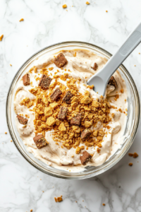 This image shows graham cracker crumbs and crushed Butterfinger candy being gently folded into the creamy peanut butter mixture using a light gray silicone spatula. The clear glass bowl sits atop a clean white marble cooktop, capturing all textures and colors of the ingredients mid-mixing.