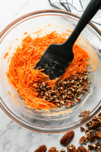 This image shows grated carrots and chopped pecans being gently folded into the mixed batter, adding texture and natural sweetness.