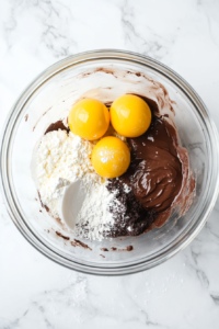 This image shows thick beaten egg yolks being folded with a cocoa, sugar, and flour mixture using a white silicone spatula in a glass bowl on a white marble countertop.