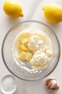 This image shows the initial ingredients for lemon garlic aioli—like crushed garlic, lemon juice, egg yolk, and oil—being added to a clear glass mixing bowl.