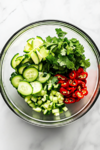 This image shows fresh cucumber rounds, diced avocado, scallions, sliced chili peppers, and cilantro leaves added to a medium mixing bowl, ready to be tossed.