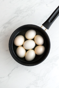 This image shows a close-up of several whole eggs resting in a saucepan, their smooth shells catching the light as they await boiling.