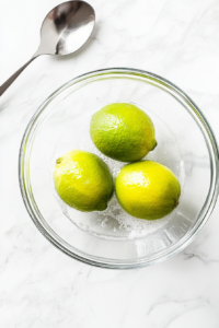 This image shows a handful of whole, glossy green limes being gently washed under running water, capturing the fresh preparation moment before making Brazilian lemonade.