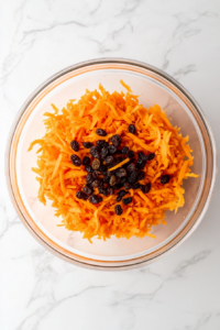 This image shows freshly shredded carrots and plump golden raisins resting in a large clear glass bowl placed on a pristine white marble cooktop.