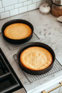 This image shows golden-brown cake layers cooling in black pans and also on a wire rack over a white marble cooktop.