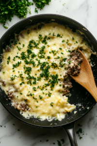 This image shows the finished cheeseburger pasta being garnished with freshly chopped parsley in a black skillet, with a wooden serving spoon resting inside, over a clean white marble countertop.