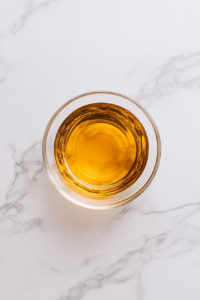 This image shows a transparent glass bowl containing oil, rum, and water, placed on a clean white marble cooktop with no background clutter.