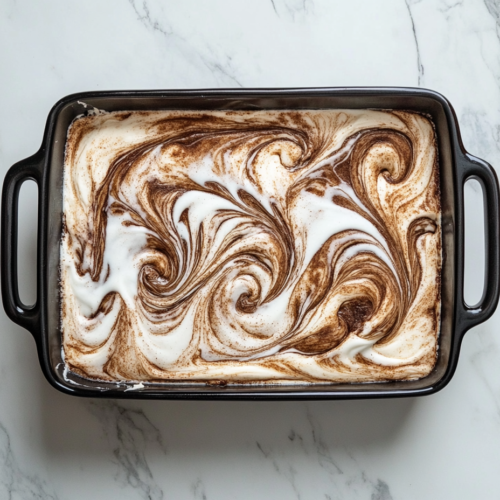 This image shows a top-down view of a baked cinnamon roll cake in a black rectangular pan, topped with glaze, over a white marble countertop.