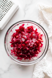This image shows freshly grated beets being placed into a clear glass bowl, forming the colorful and hearty base for a beetroot salad.