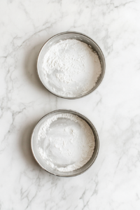 This image shows two greased and floured 9-inch aluminum baking pans ready for the chocolate cake batter, placed on a white marble countertop.