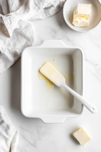 This image shows a hand greasing a 9x9 inch baking dish while the oven is being preheated, setting the stage for a delicious baked pineapple dessert.