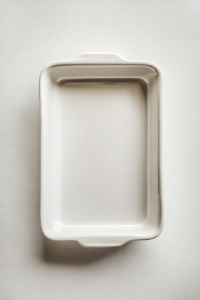 This image shows a clean, white 9x5-inch loaf pan being greased with golden olive oil using a silicone pastry brush. The pan sits on a white marble cooktop under natural lighting, with the olive oil bottle neatly placed beside it. The background is completely clear, keeping full focus on the preparation process.